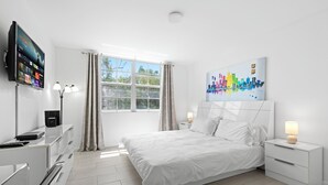 A modern bedroom with a bed, bedside tables, a TV, and a window with curtains. - Yacht Club Miami Suite 2 Bedroom and 2 Bathroom With Baby Crib & Free Parking (Aventura)