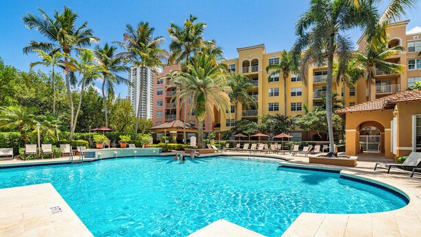 A swimming pool with lounge chairs and palm trees, surrounded by multi-story buildings. - Yacht Club Miami Suite 2 Bedroom and 2 Bathroom With Baby Crib & Free Parking (Aventura)