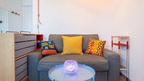 Ein modernes Wohnzimmer mit einem grauen Sofa, bunten Kissen, einem kleinen blauen Tisch mit einem violetten Gegenstand und einer Holzkombination mit Schubladen.