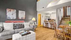 Uma sala de estar moderna com um sofá cinzento, uma mesa de jantar de madeira com cadeiras e uma escadaria que dá para um andar superior.