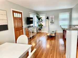 A modern kitchen with a granite countertop, a dining area with a white table, and a living space with a TV and plants.