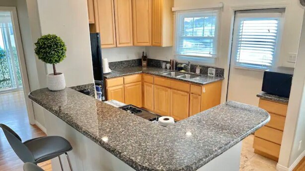 A kitchen with granite countertops, wooden cabinets, a central island with bar stools, and a potted plant. - RustlingLeaves.Kg&Qn.2ndFlrGarageLaundry (Los Angeles)