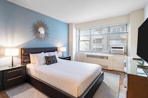 A modern bedroom with a large bed, bedside lamps, a decorative mirror, and a window with blinds.