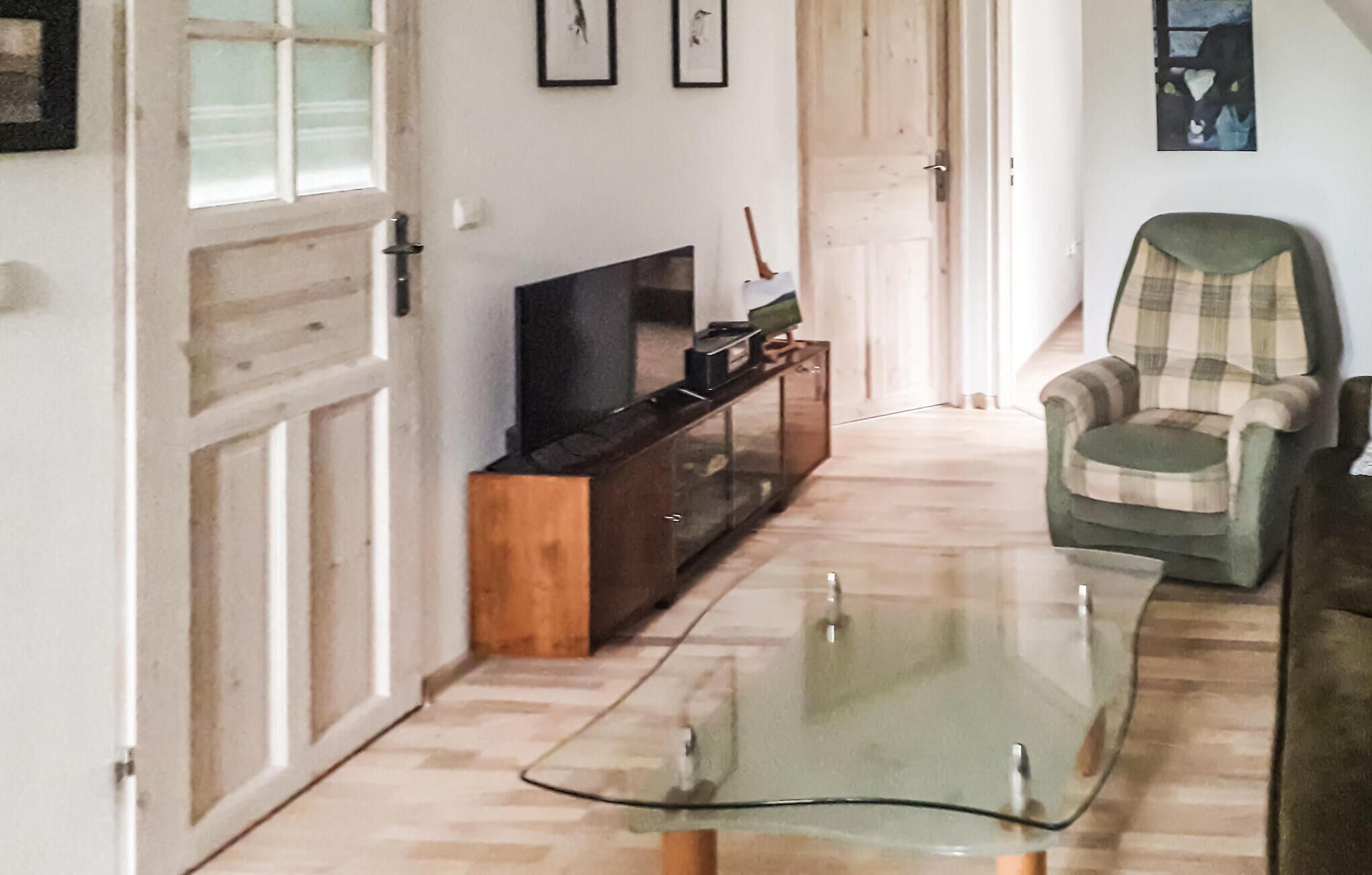 A living room with a glass coffee table, a television on a wooden stand, a plaid armchair, and a door leading to another room.