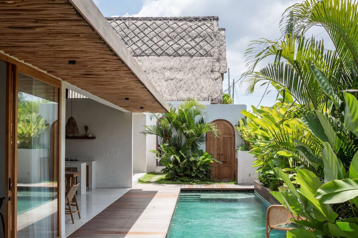 A modern outdoor pool area with a wooden deck, a glass door entrance, and lush tropical plants.
