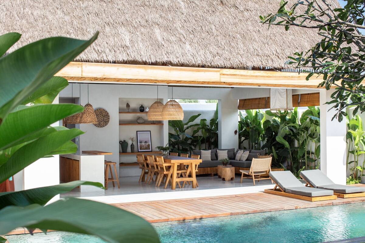 A poolside area with a thatched roof, wooden furniture, and lush greenery.