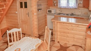 Eine gemütliche Holzhütte mit Küchenbereich, inklusive Spüle, Mikrowelle und kleinem Kühlschrank. Ein Esstisch mit Stühlen und einer Tischdecke aus Spitze ist ebenfalls vorhanden.