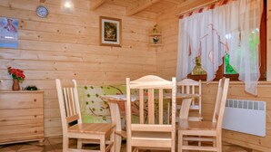 Ein Holzhütten-Zimmer mit einem Bett, zwei Holzstühlen, einem Schminktisch, einer Uhr, einem gerahmten Bild und einem Fenster mit Vorhängen.