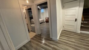 Un couloir doté d’un plancher en bois, de murs blancs et d’une porte donnant sur une chambre à coucher, où se trouvent un lit et une fenêtre.