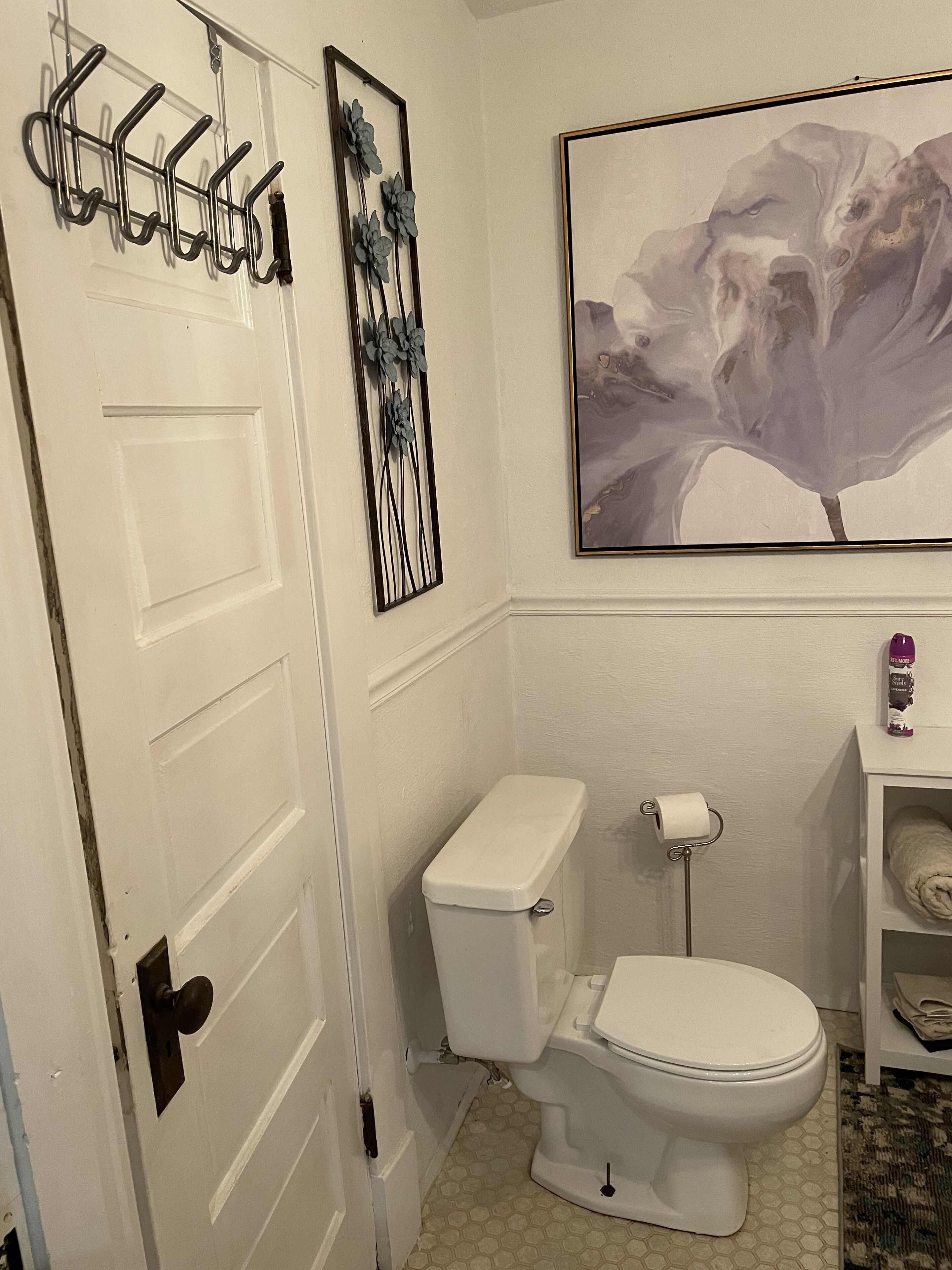 Un baño con inodoro, soporte para papel higiénico, toallero, cuadro abstracto enmarco y estante con una toalla enrollada.