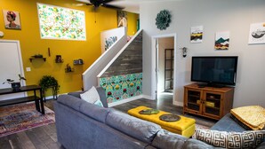 Una sala de estar con un sofá gris, un otoman amarillo, una escalera con una pared colorida, un televisor y un mueble de madera.