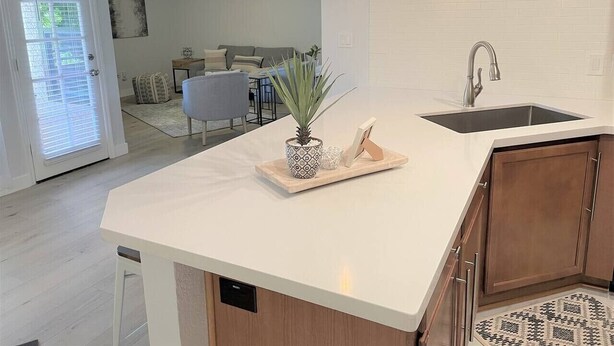 Uma cozinha moderna com bancada branca, armários de madeira, pia embutida, iluminação pendente e um vaso com planta.