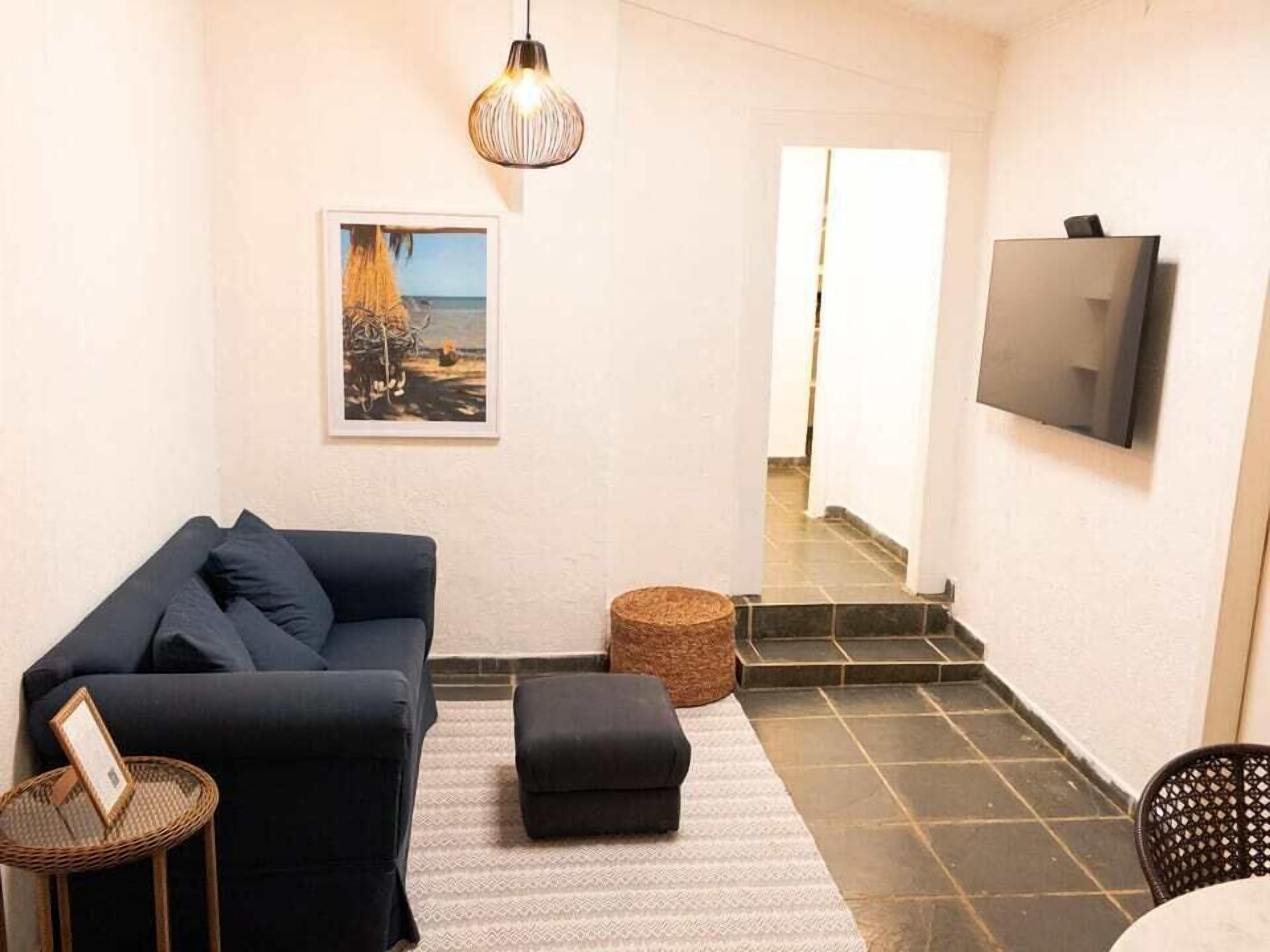 A living room with a dark blue sofa, a small round ottoman, a wicker side table, a framed picture on the wall, a hanging light fixture, and a TV mounted on the wall.