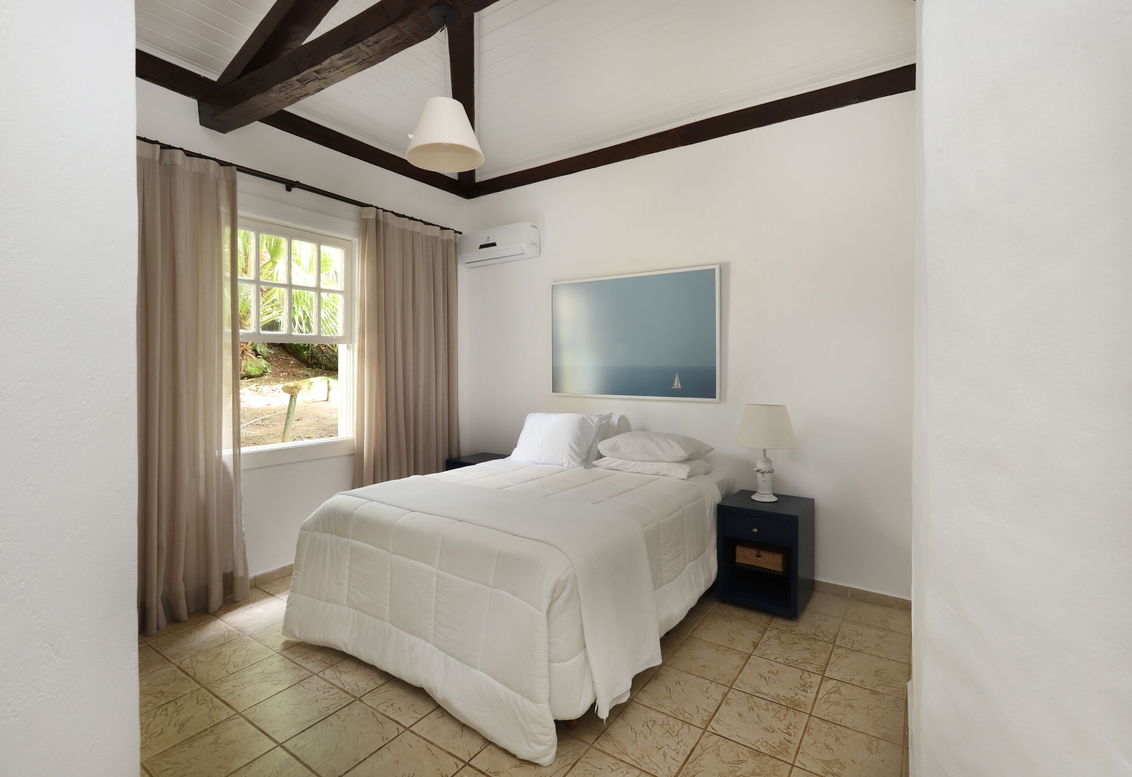 A bedroom with a bed, a nightstand, a lamp, a window with curtains, and a painting on the wall.
