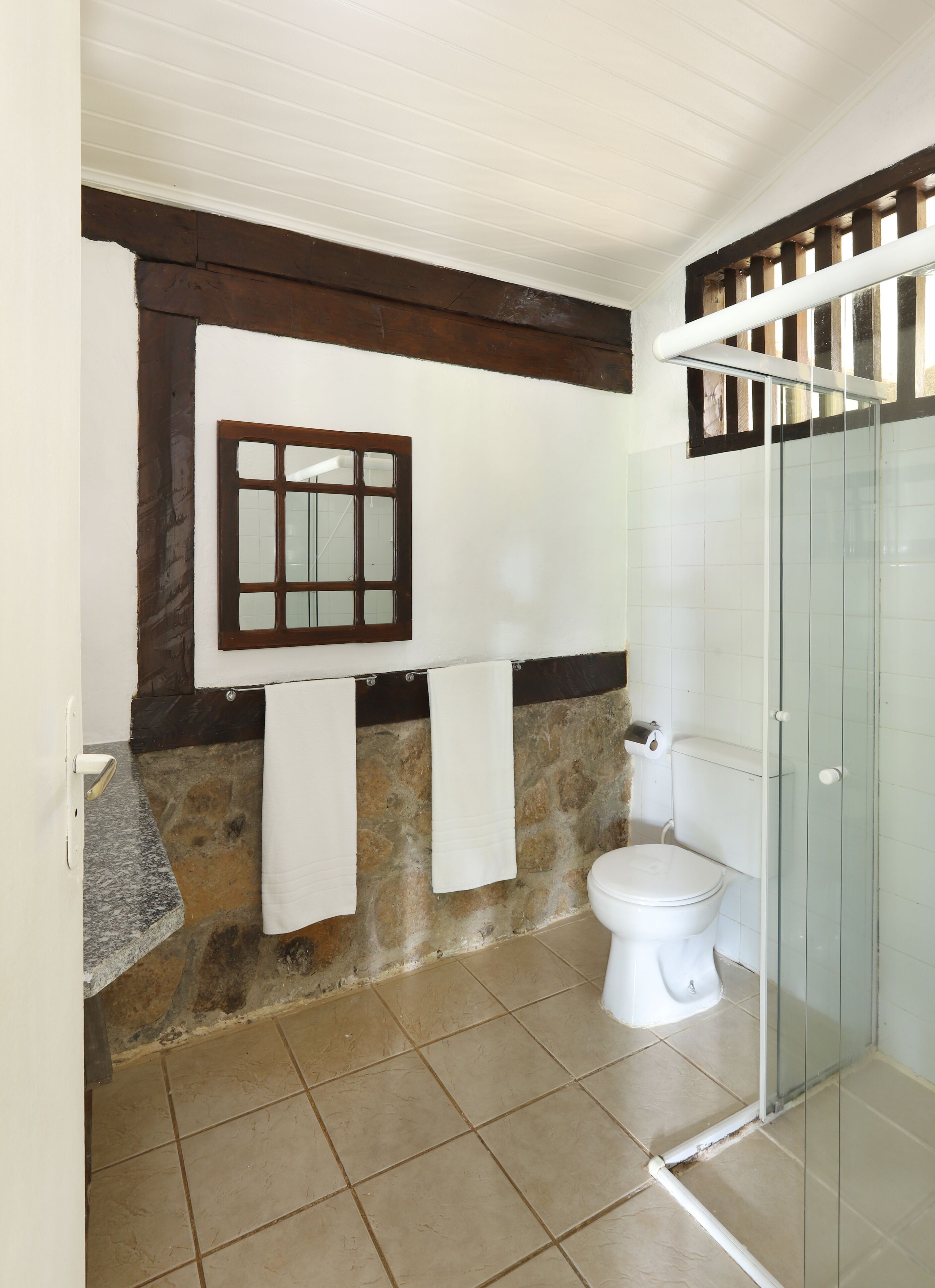 A bathroom with a stone wall, a white toilet, a glass shower enclosure, and a window with a wooden frame.