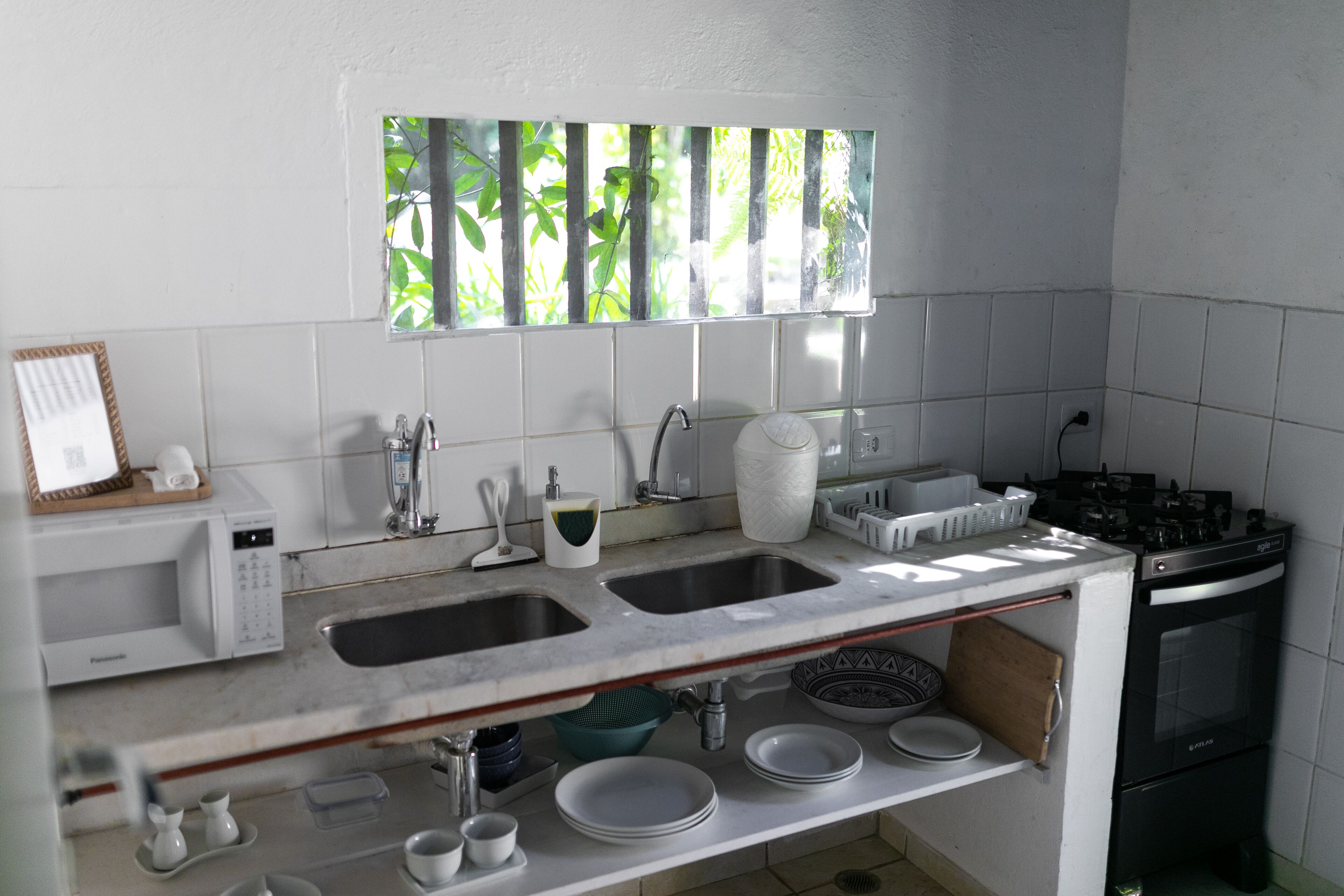 A kitchen with white tiles, a window with a view of greenery, a microwave, a sink, and an oven.