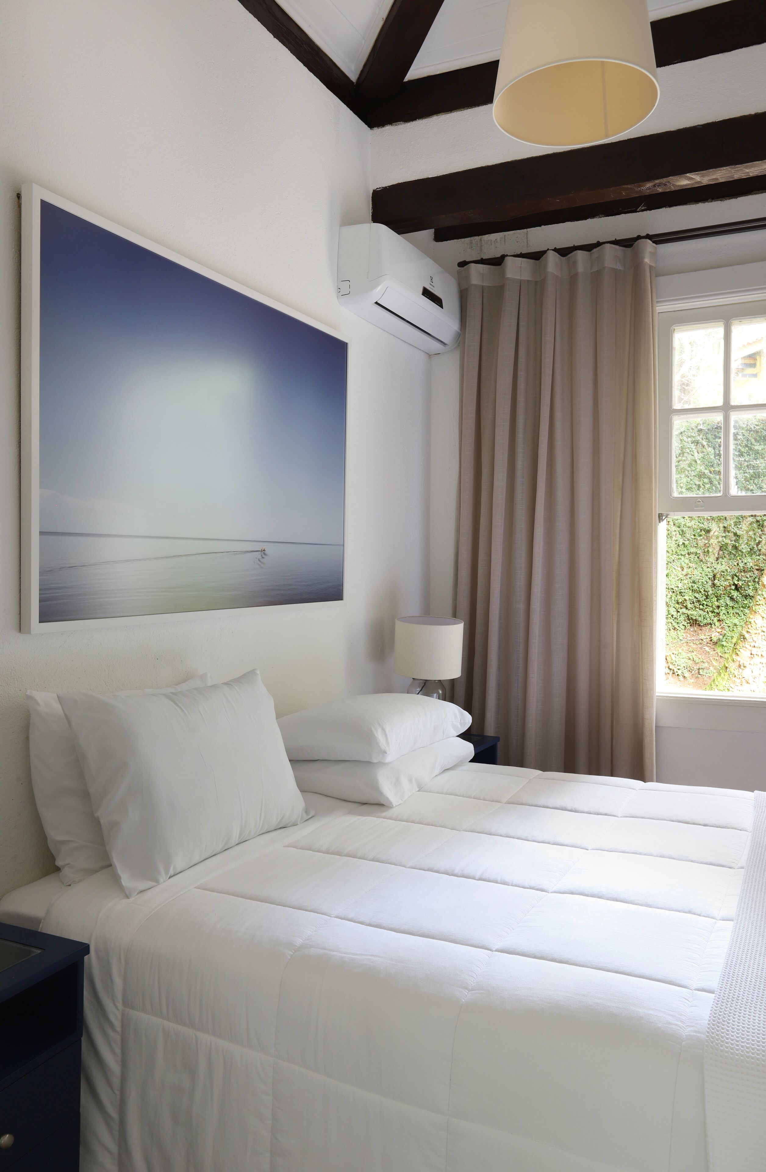 A neatly made bed with white bedding, a bedside lamp, and a large painting of a beach scene on the wall.