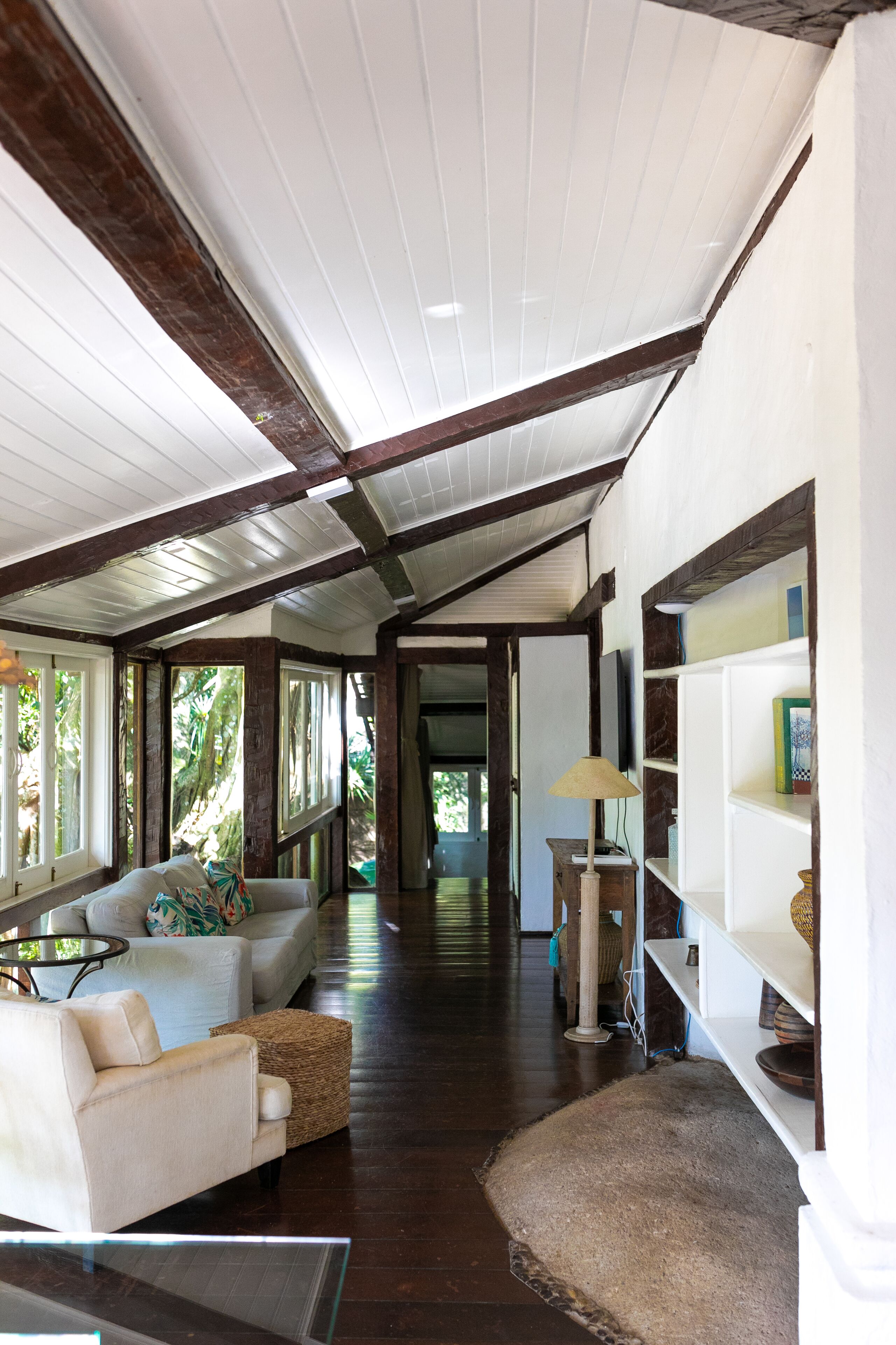 A spacious hallway with a high ceiling, wooden beams, a white sofa, a wicker ottoman, a glass coffee table, and a bookshelf.