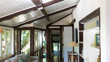 A spacious hallway with a high ceiling, wooden beams, a white sofa, a wicker ottoman, a glass coffee table, and a bookshelf.