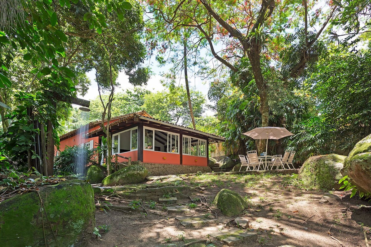 Een huis met een rode gevel en grote ramen, omgeven door weelderige begroeiing en rotsen.