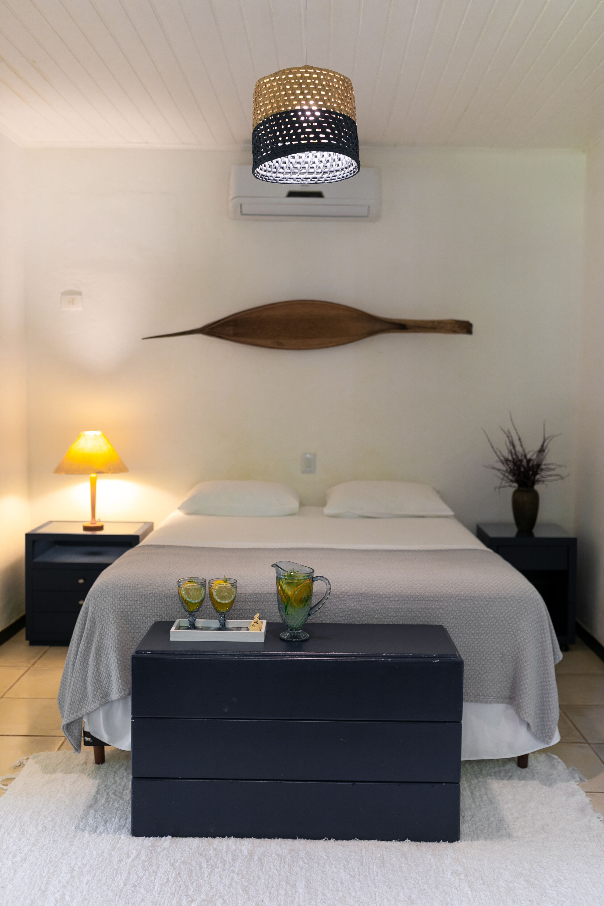 A bedroom with a bed, bedside tables, a lamp, a decorative wall piece, a vase, and a small table with glasses and a pitcher.