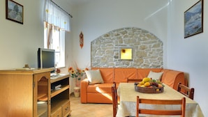 A cozy living room with a stone wall, orange sofa, wooden dining table with a fruit basket, and a TV on a wooden cabinet. - 1 bedroom beach front apartment in Rakalj (Rakalj)