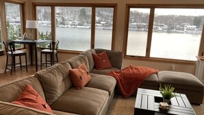 A living room with a large window overlooking a lake, a sofa, a coffee table, and a dining area with chairs. - Enjoy Lakefront Sunsets in Cozy 2BR Cottage Escape (Plymouth)