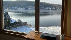 A view of a lake with snow-covered ground and trees, seen through a window with a wooden table and a clock. - Enjoy Lakefront Sunsets in Cozy 2BR Cottage Escape (Plymouth)