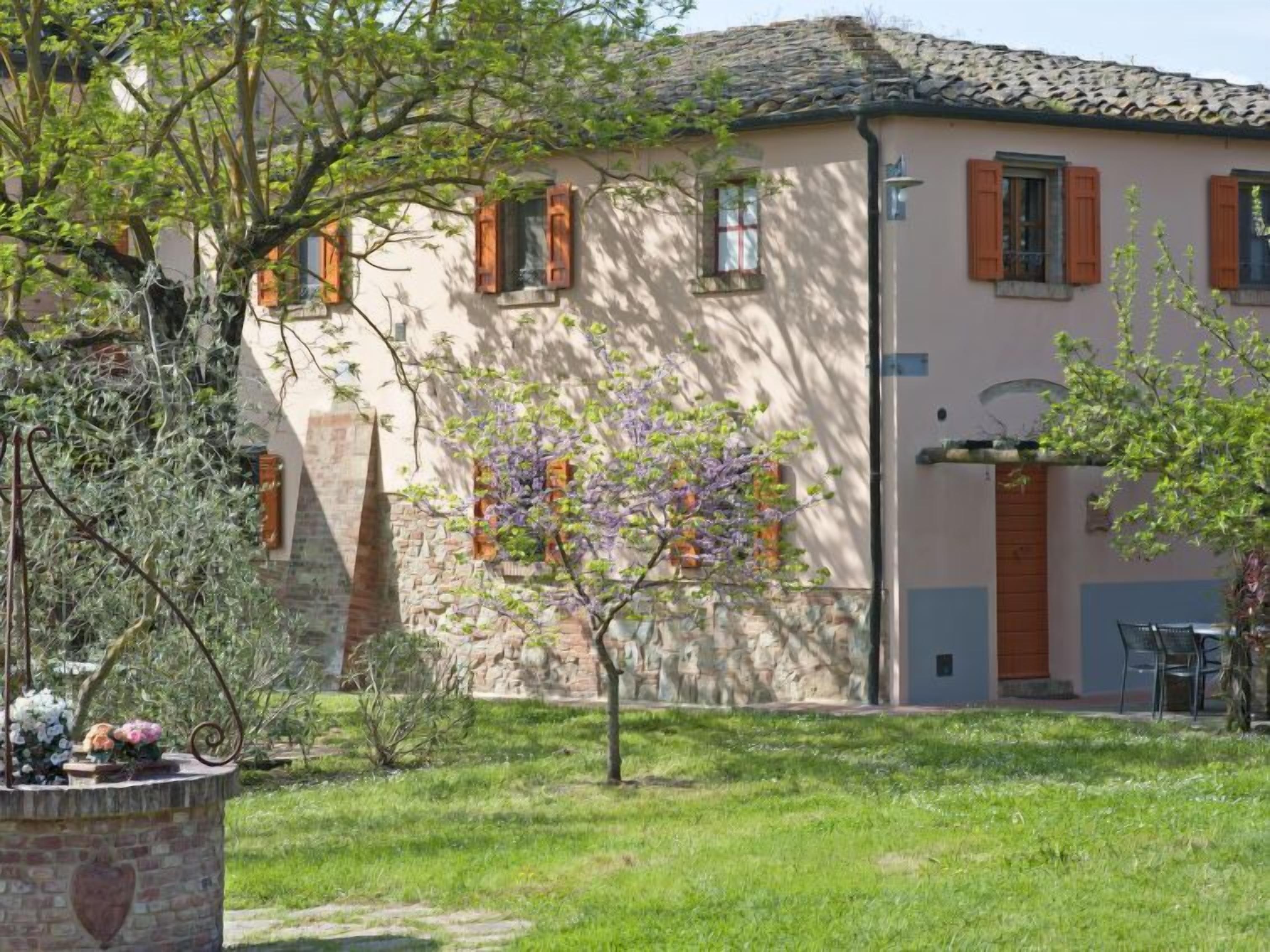 A traditional building with a stone facade, wooden shutters, and a small garden area with a tree and a flower arrangement.