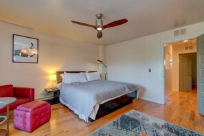 Une chambre à coucher avec un lit, un fauteuil rouge, une table de chevet, une lampe, un ventilateur de plafond et une porte donnant sur une autre pièce.