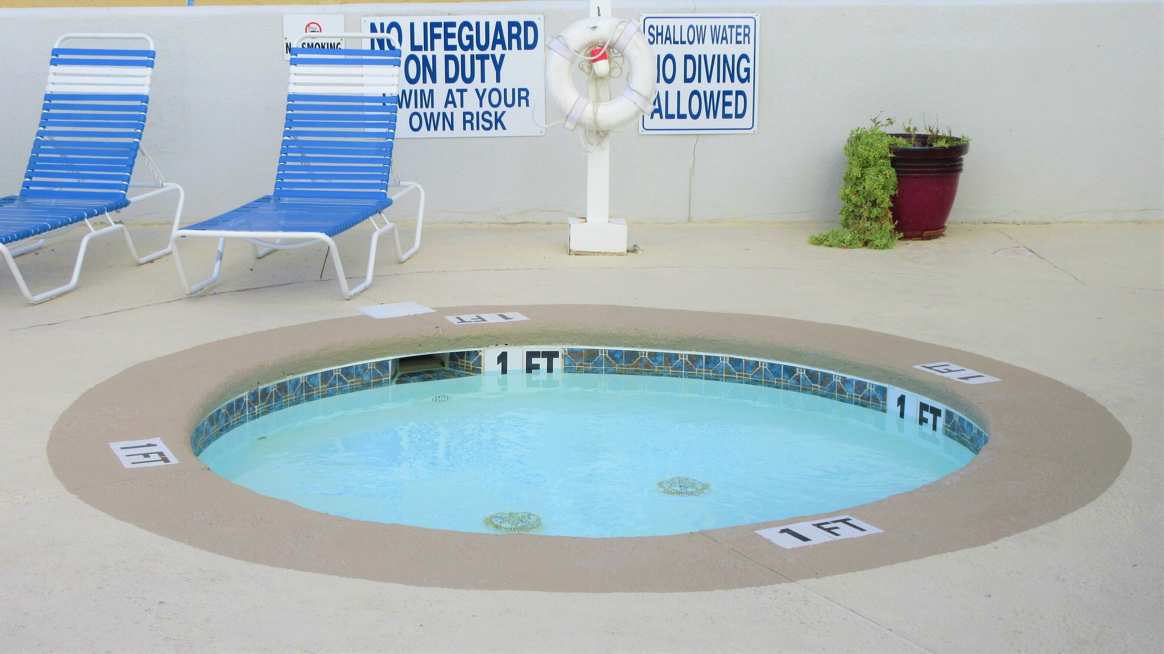 A small pool with a 1-foot depth marker, lounge chairs, and warning signs about shallow water and no lifeguard on duty.