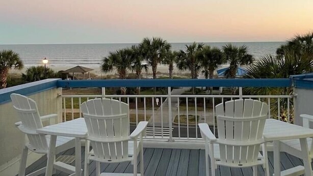 Een houten terras met witte stoelen en uitzicht op de oceaan bij zonsondergang.
