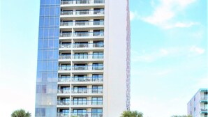 Een hoog, modern hotel met meerdere balkons, een overdekte doorgang en palmbomen.