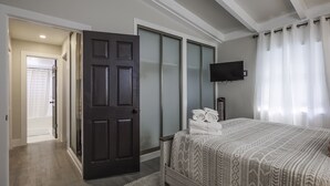 Ein Schlafzimmer mit einem Bett, einer dunklen Tür, einem an der Wand befestigten Fernseher und einem Fenster mit Vorhängen.