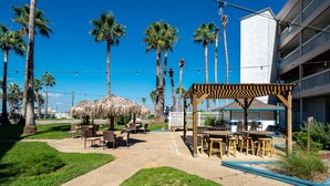 An outdoor patio area with thatched umbrellas, wooden tables, and chairs, surrounded by palm trees and a building with balconies. - Sunrise Beach Retreat (Corpus Christi)