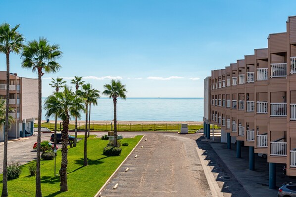 A hotel with a view of the sea, palm trees, and a parking lot.