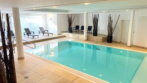 Una amplia piscina interior con suelo de baldosas, sillas de descanso y altas plantas decorativas.