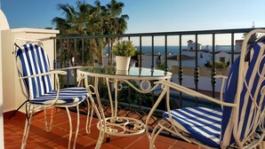 Un balcon avec deux chaises à rayures et une table, offrant une vue sur la côte, avec des palmiers et des bâtiments.