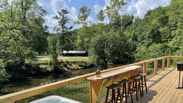 A wooden deck with a hot tub, bar stools, and a view of a river and trees.