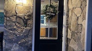 A black front door with a stone exterior, a welcome wreath, and a small step leading to the entrance.