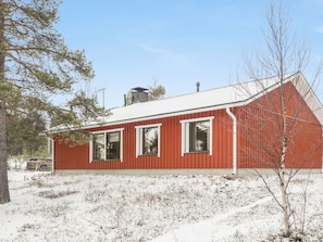 A red wooden house with a white roof, surrounded by snow-covered ground and sparse vegetation.