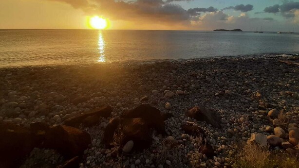 A pebbled beach with the sun setting over the water, boats in the distance, and a cloudy sky.