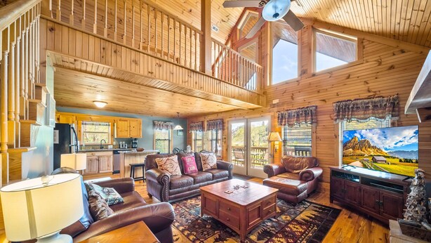 A spacious living room with a high ceiling, wooden beams, a fireplace, and a large TV. - Cruizin River Dreams- Riverfront, fire pit, hot tub (Todd)
