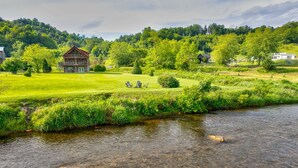 A riverside view with a wooden cabin, a grassy field, and a forested hill. - Cruizin River Dreams- Riverfront, fire pit, hot tub (Todd)