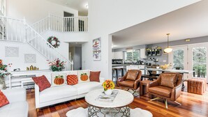 A spacious living area with a white sofa, brown leather chairs, and a round coffee table. A staircase with a railing leads to an upper level. - Sterling Valley Home ~ 9 Mi to Stowe Mountain! (Morristown)
