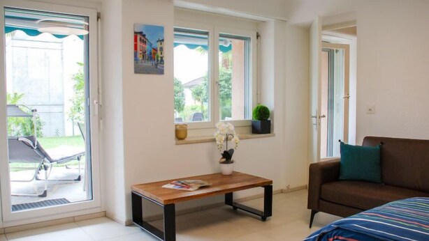 A room with a bed, a sofa, a table, and a window with a view of a house and greenery. - Appartamento 2- Giardino by Interhome (Locarno)