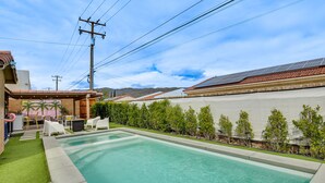 A rectangular swimming pool with a tiled deck, surrounded by a well-maintained hedge and a patio area with outdoor furniture. - Desert Hot Springs Home w/ Pool & Gas Grill! (Desert Hot Springs)
