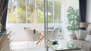 A modern living room with a glass coffee table, wicker chairs, a sofa, and a view of trees through large windows.