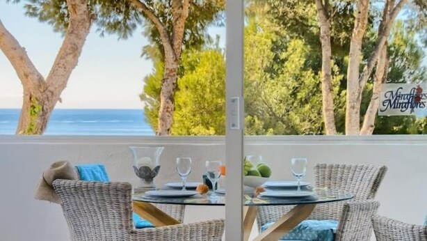 A dining area with wicker chairs and a glass table, overlooking a beach with trees and a sign reading "Miraflores Minifloras".