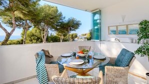 A balcony with a glass table set for dining, wicker chairs, and a view of trees and the sea.